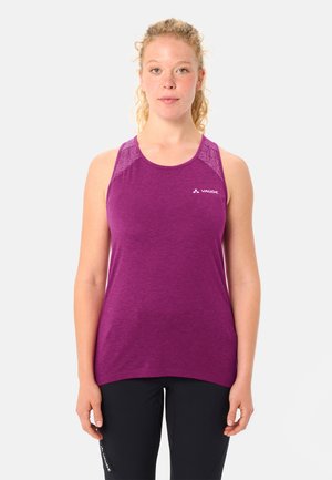 Frau mit lockigem blondem Haar, die ein ärmelloses lila Sportoberteil und schwarze Leggings trägt, vor einem einfarbigen hellen Hintergrund stehend.