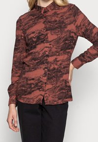Camicia a maniche lunghe color ruggine con un motivo marmorizzato nero, chiusura frontale con bottoni e colletto. Realizzata in tessuto morbido con una texture liscia.
