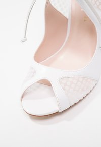 Chaussure blanche à talon haut ouvert à l'avant avec des panneaux en mesh à pois et une bride de cheville à lacets, vue en gros plan de face.