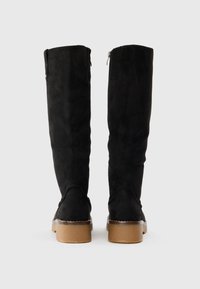 Bottes noir en daim hautes jusqu'au genou avec une texture lisse, fermeture éclair sur le côté et semelle en caoutchouc marron contrastante. Design épuré et minimaliste.