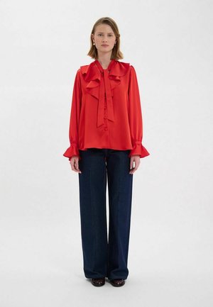 Blouse en satin rouge avec col et manches volantés, présentant une coupe ample et un nœud à l'avant. Associée à un pantalon en denim bleu foncé à jambes larges.