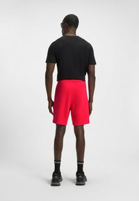 Pantaloni corti rossi con una tasca sul retro, indossati con una maglietta nera. Il tessuto appare liscio e la vestibilità è comoda. Scarpe da ginnastica nere e calzini sono visibili.