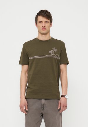 Άνδρας όρθιος που φοράει ελιάς πράσινο T-shirt με σχεδιασμό φοίνικα και ήλιου σε γραμμή, γκρι παντελόνια και καφέ ρολόι χειρός στον αριστερό καρπό.