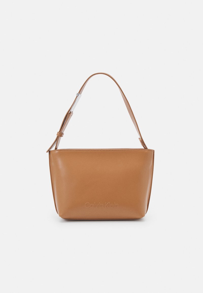 Tan nahkalaukku, jossa on sileä pinta, suorakulmainen muoto, säädettävä hihna ja kohokuvioitu "Calvin Klein" -logo edessä.
