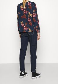 Blusa floreale blu navy con motivi rossi e gialli, abbinata a jeans scuri e sneakers alte nere. Maniche lunghe con polsini elasticizzati.