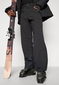 Svarta skidbyxor med rak passform, utrustade med fickor med dragkedja och förstärkt tyg vid fållarna, i kombination med svarta skidpjäxor och en snowboard.