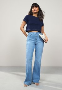 T-shirt marine court, jean évasé bleu clair, sac à main blanc et sandales de couleur claire. Le tissu est lisse ; le jean présente une taille haute et une décoloration.