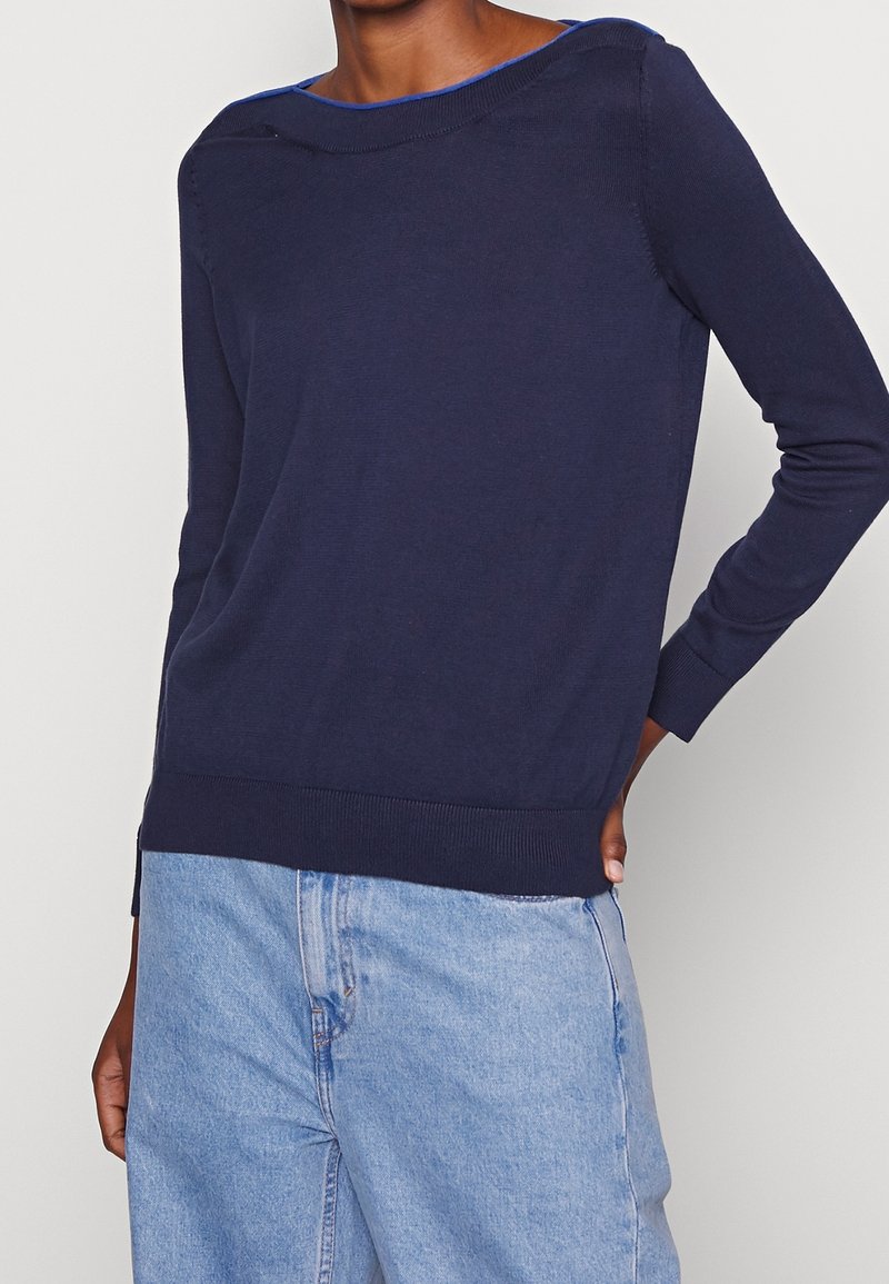 Personne portant un pull à manches longues bleu marine et un jean taille haute bleu clair, debout avec une main posée sur la hanche.