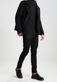 Un cappotto doppiopetto nero sopra un maglione nero testurizzato, abbinato a pantaloni neri slim-fit e scarpe stringate nere, creando un look monocromatico.