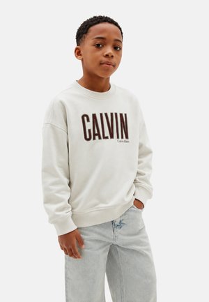 Ragazzo che indossa una felpa bianca Calvin Klein e jeans azzurri, in piedi con una mano in tasca davanti a uno sfondo semplice.