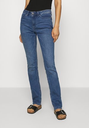 Personne portant un jean slim bleu, un haut noir et des sandales slides noires avec une grande boucle, debout devant un fond uni.