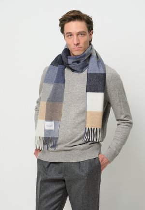 Wollen sjaal in een blauw, grijs en beige ruitpatroon, met franjes aan de randen en een logo-label. Gedragen over een grijze trui.