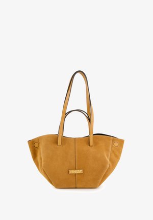 Sac fourre-tout en daim couleur tan avec deux longues poignées et une couture centrale. Présente une plaque de marque et des accents circulaires. Fini texturé et doux.