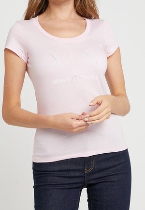 Femme portant un t-shirt à manches courtes rose clair Armani Exchange avec un logo "A|X" en strass et un jean bleu foncé, les mains croisées devant.