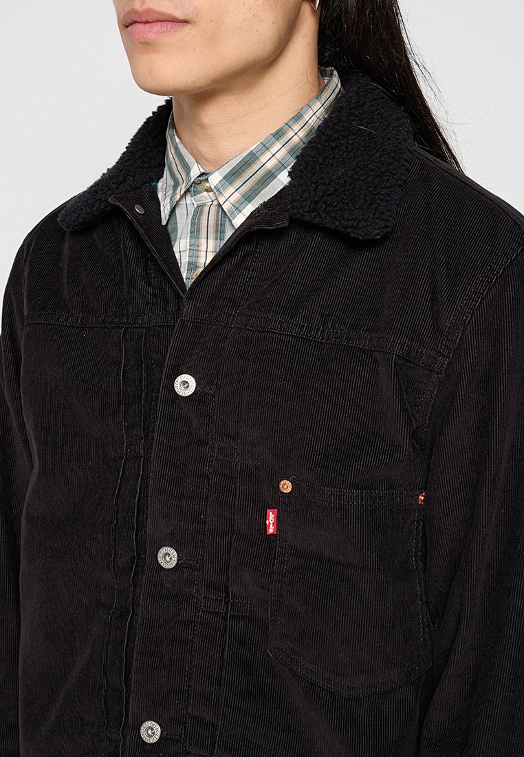 Chaqueta de pana negra con un cuello texturizado y esponjoso, que cuenta con cierres de botón, un bolsillo en el pecho y el detalle de la etiqueta roja de Levi's.
