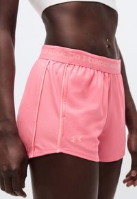 Κοντινό πλάνο ενός ατόμου που φοράει ροζ αθλητικά σορτς Under Armour και λευκό αθλητικό σουτιέν, με εμφανή γυμνασμένη κοιλιακή περιοχή και σκουλαρίκια στον αφαλό.