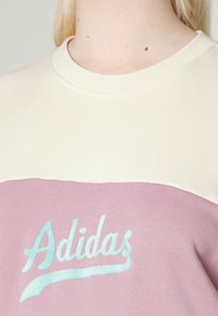 Felpa di cotone con una parte superiore color crema e una parte inferiore rosa. Presenta un logo "Adidas" azzurro chiaro in corsivo sulla parte anteriore.
