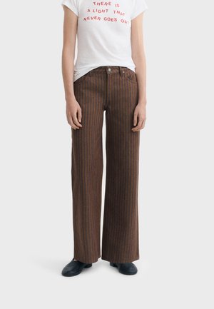 Personne portant un pantalon large marron à fines rayures, des chaussures noires et un t-shirt blanc avec un texte rouge disant "There is a light that never goes out."