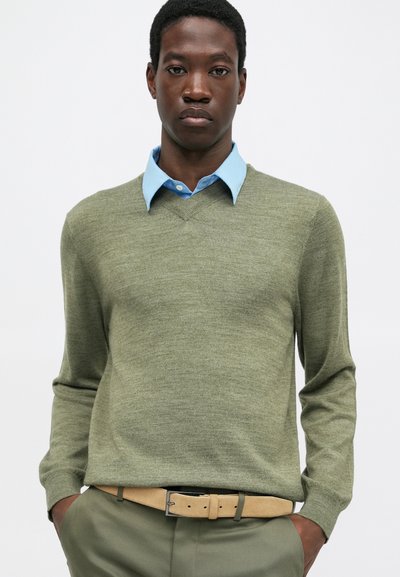 Giovane uomo con una camicia azzurro chiaro sotto un maglione verde oliva a V, abbinato a pantaloni verde oliva e una cintura beige.