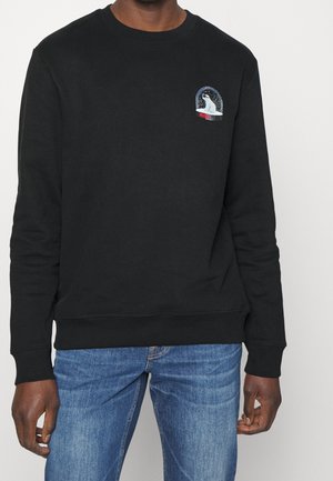 Schwarzer Pullover mit langen Ärmeln und kleinem weißem Eisbär in einem Schneekugel-Design auf der linken Brust, getragen mit blauen Jeans.