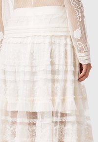 Λευκή φούστα από δαντέλα με πολυεπίπεδα βολάν, διακοσμημένη με κεντήματα λουλουδιών και διάφανο ύφασμα. Το υφή υφάσματος δημιουργεί εμφάνιση ανάλαφρη και αέρινη.