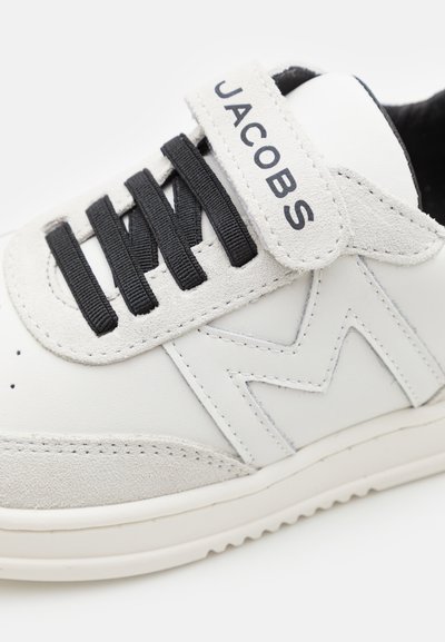 Witte leren sneaker met grijze suède accenten, zwarte veters en een grijze band met de tekst "JACOBS". Kenmerkend zigzagontwerp aan de zijkant.
