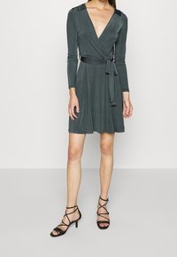 Vestito a portafoglio grigio scuro, con scollatura a V, maniche lunghe e cintura da annodare. Tessuto morbido con una silhouette fluida. Tacchi neri con cinturini.