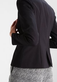 Person, die ein tailliertes schwarzes Blazer und schwarz-weiße gepunktete Hose trägt und das Blazer von hinten anpasst.