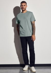 Svetle zelené tričko s krátkým rukávem a kulatým výstřihem, v kombinaci s černými slim-fit kalhotami a off-white teniskami. Model stojí proti neutrálnímu pozadí.