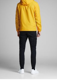 Žlutá nepromokavá bunda s kapucí, kombinovaná s černými cargo kalhotami a bílými teniskami. Bunda má hladkou texturu a je bez ozdob.