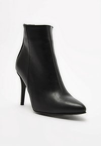 Bottine en cuir noir avec un bout pointu et un talon stiletto haut. Texture lisse avec un design minimaliste et une fermeture éclair à l'arrière.