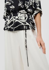 Zwarte blouse met wit bloemenpatroon, losse pasvorm, getailleerd met een trekkoord, gecombineerd met vloeiende witte broeken. Soepele textuur.