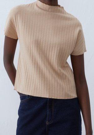 Personne portant un haut beige côtelé à manches courtes associé à une jupe en denim bleu foncé, debout devant un fond clair.
