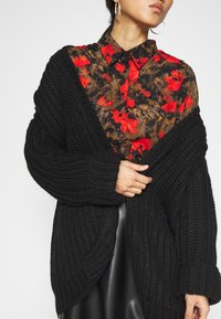 Cárdigan negro de punto con un grueso ribete. Lleva puesto sobre una camisa de estampado floral con tonos rojos y marrones. Manga larga y apertura frontal.