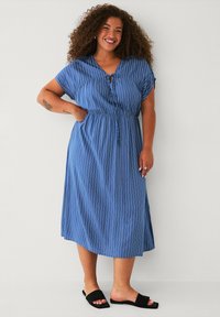 Abito blu a righe con maniche corte, dettagli con bottoni, vita elastica e orlo fino al ginocchio. Abbinato a sandali neri a ciabatta.