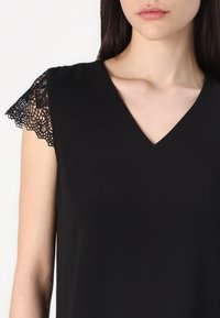 Sort V-hals bluse med blondekantede halværmer. Glat tekstur, tætsiddende design og delikat blodedetalje på skuldrene.
