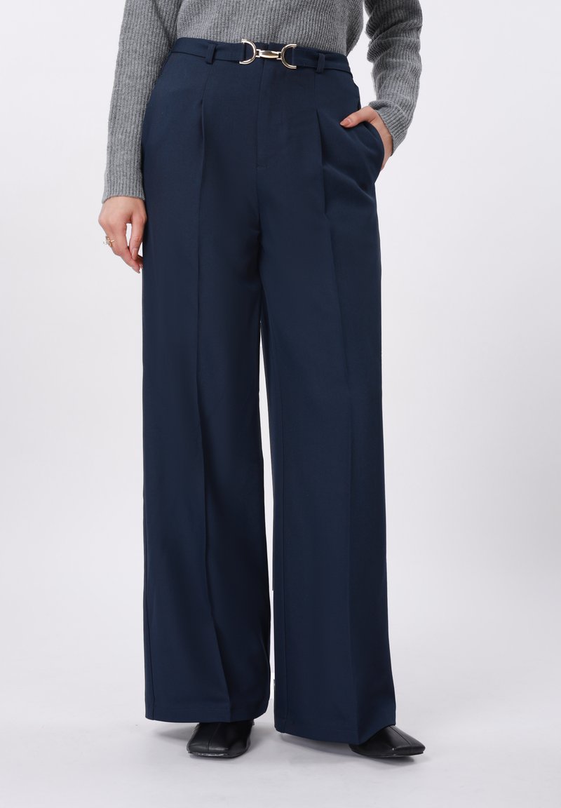 Pantalon large marine à taille haute, avec plis sur le devant et boucle de ceinture double anneau. Le tissu paraît lisse avec une coupe sur mesure.
