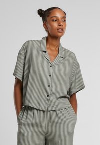 Cropped short-sleeve shirt in olive green with white vertical stripes, featuring a notched collar and black buttons. Soft, smooth fabric.