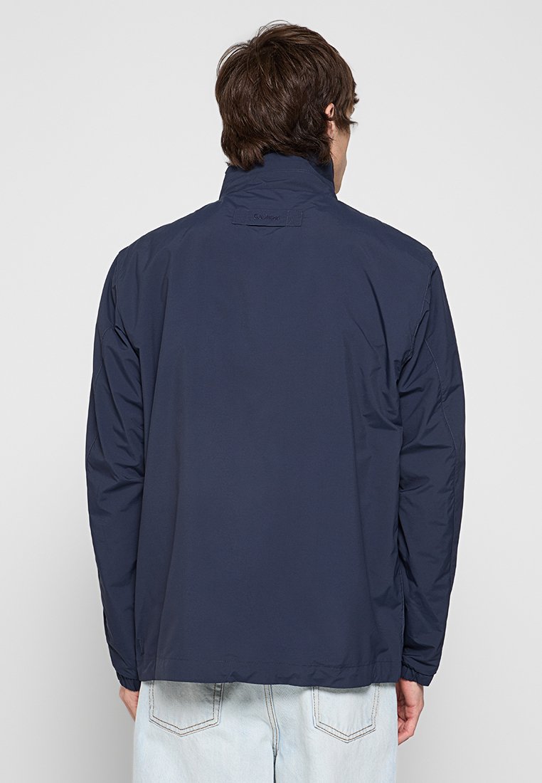 Marineblaue leichtgewichtige Jacke mit hohem Kragen, strukturiertem Stoff und dezentem Logo auf der Rückseite. Entworfen für Komfort und Bewegungsfreiheit.