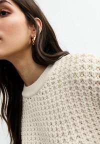 Pull en laine de couleur crème avec un motif texturé, col ribbée, porté avec une boucle d'oreille en or, visage partiellement visible et cheveux bruns.