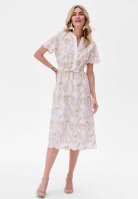 Witte jurk met gouden bloemenpatroon, korte mouwen en een V-hals. Elastische taille en golvende zoom. Het model draagt beige ballerina's en oorbellen.