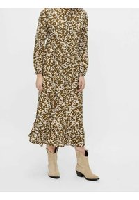 Kvinna klädd i en brun blommig midi-klänning med långa ärmar och beige ankel-kowboystövlar, stående mot en enfärgad bakgrund.