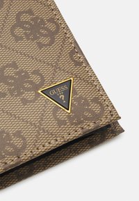 Brown patterned wallet with a textured synthetic material. Features a gold triangle badge with "GUESS USA" in black.