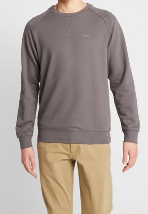 Mand iført grå langærmet sweatshirt med "s.Oliver" logo på brystet og beige bukser, stående mod en ensfarvet grå baggrund.