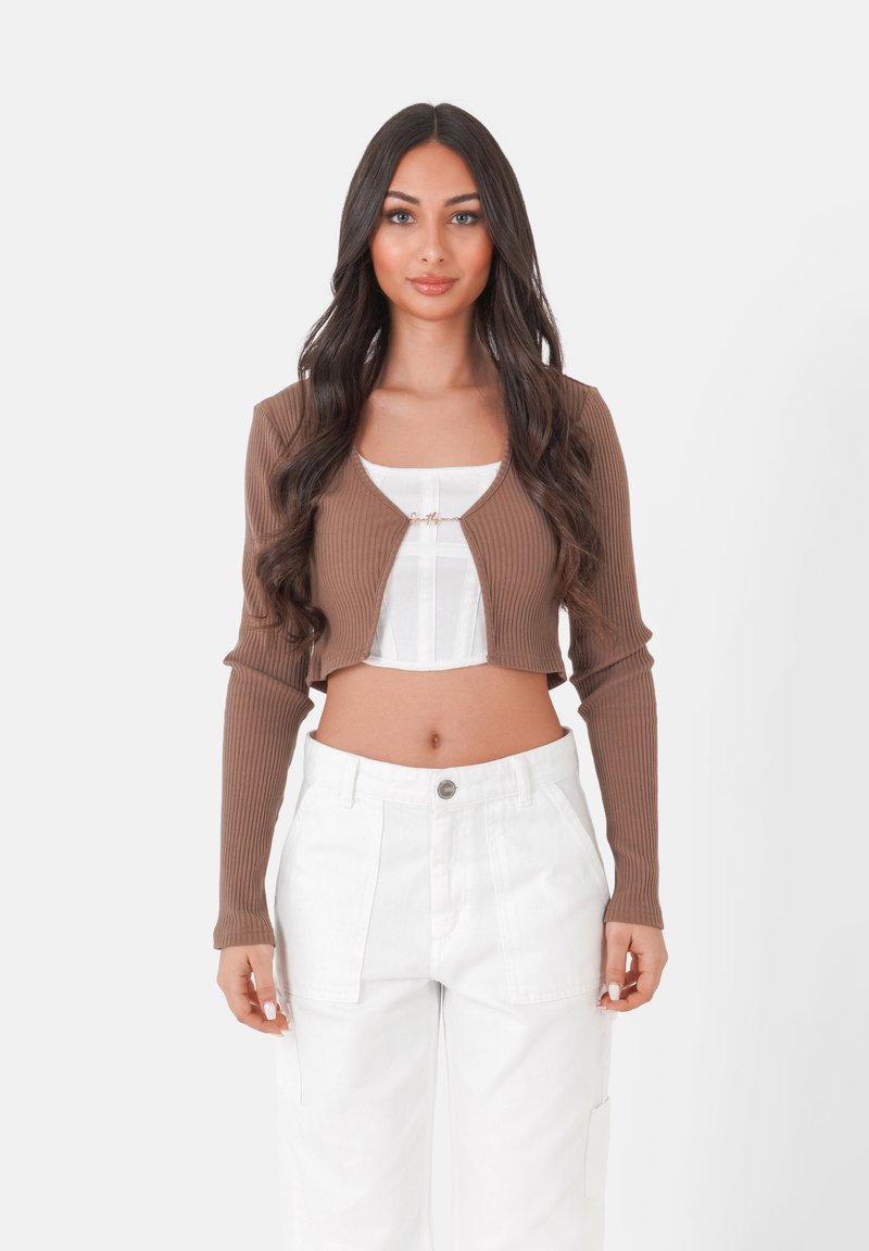 Cardigan court côtelé marron à manches longues, avec une fermeture à l'avant et un top blanc superposé en dessous, associé à un pantalon large blanc.