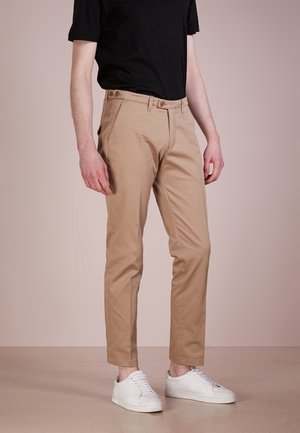 Mann trägt beige Hose, schwarzes Hemd und weiße Sneaker und steht vor einem schlichten hellen Hintergrund.