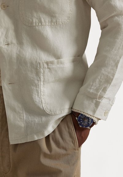 Light beige linen shirt with a pocket, paired with tan pants. A blue watch is visible on the wrist, contrasting the shirt's color.