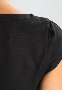 Sort top med korte ærmer, der har en tekstureret overflade og en let glans. Udskæringen har en diskret rundet form.