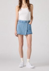 Mini jupe en denim bleu clair avec un design noué, poches avant et un bouton supérieur. Associée à un débardeur blanc et des baskets blanches.