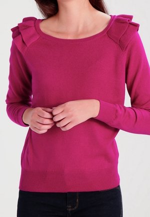 Kvinde iført en langærmet magentarød sweater med flæsedetaljer på skuldrene, kombineret med mørke bukser, hænderne løst samlet ved taljen.